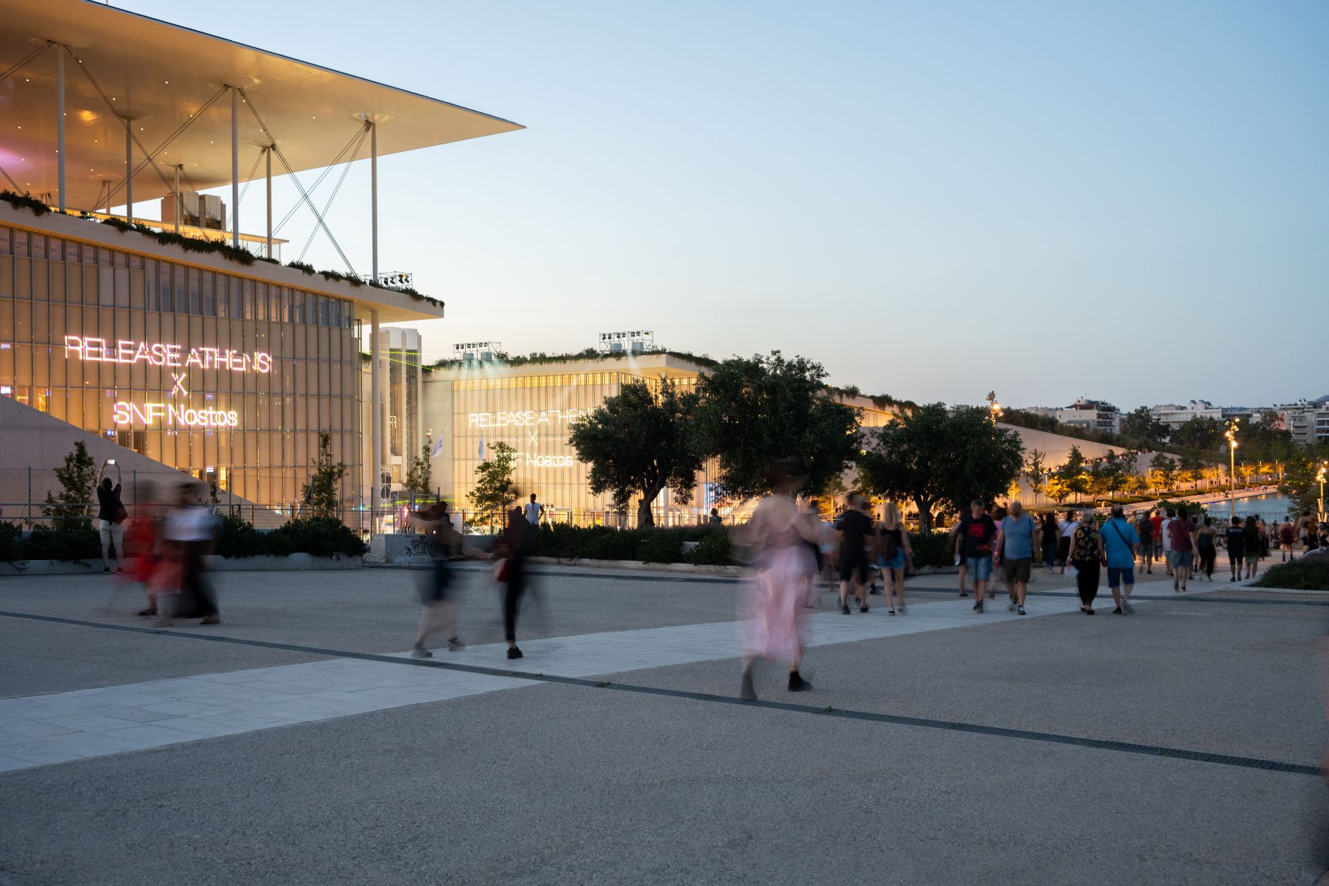 Εξωτερικό ΚΠΙΣΝ το σούρουπο με «Release Athens x SNF Nostos» προβαλλόμενο στη γυάλινη πρόσοψη. Θολές φιγούρες στην πλατεία. 